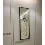 مرآة جدارية مستطيلة بإطار أسود وزخارف نباتية بيضاء، تضيف لمسة عصرية للأماكن.