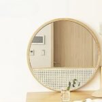 مرآة جدارية دائرية بتصميم عصري، إطار خشبي فاتح مع قاعدة مزخرفة باللون الأبيض والأخضر.