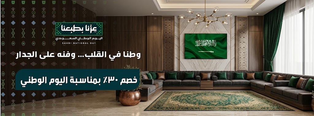 أريكة كبيرة بلون رمادي داكن مع وسائد خضراء، وسجادة مزخرفة، وتلفاز يعرض العلم السعودي في ديكور عصري.